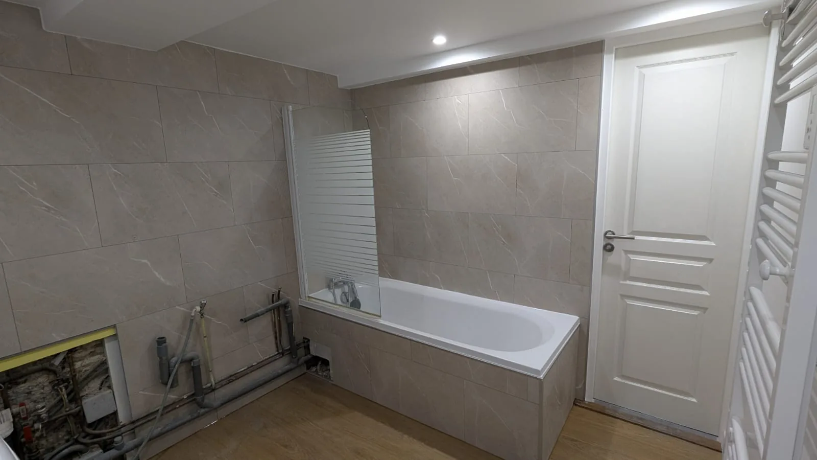 Salle de bains avec baignoire carrelée, sol imitation bois et raccordements visibles le long du mur.