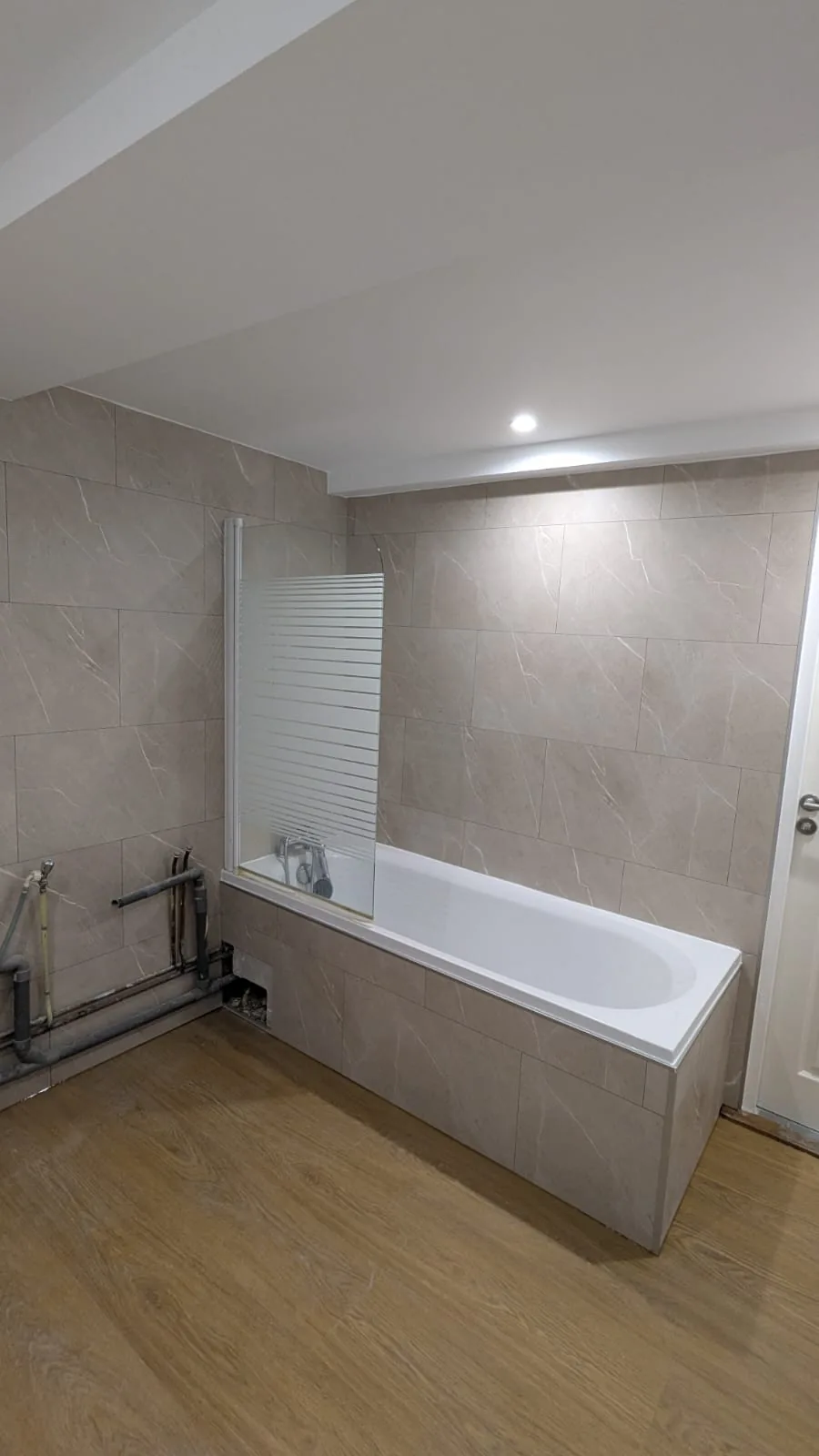 Baignoire habillée de carreaux beige au-dessus d'un sol imitation bois, avec raccordements apparents.