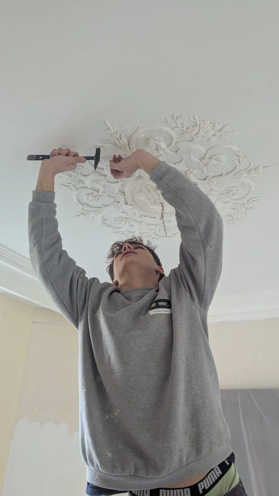 Artisan vu en contre-plongée intervenant sur une grande rosace florale blanche au plafond.