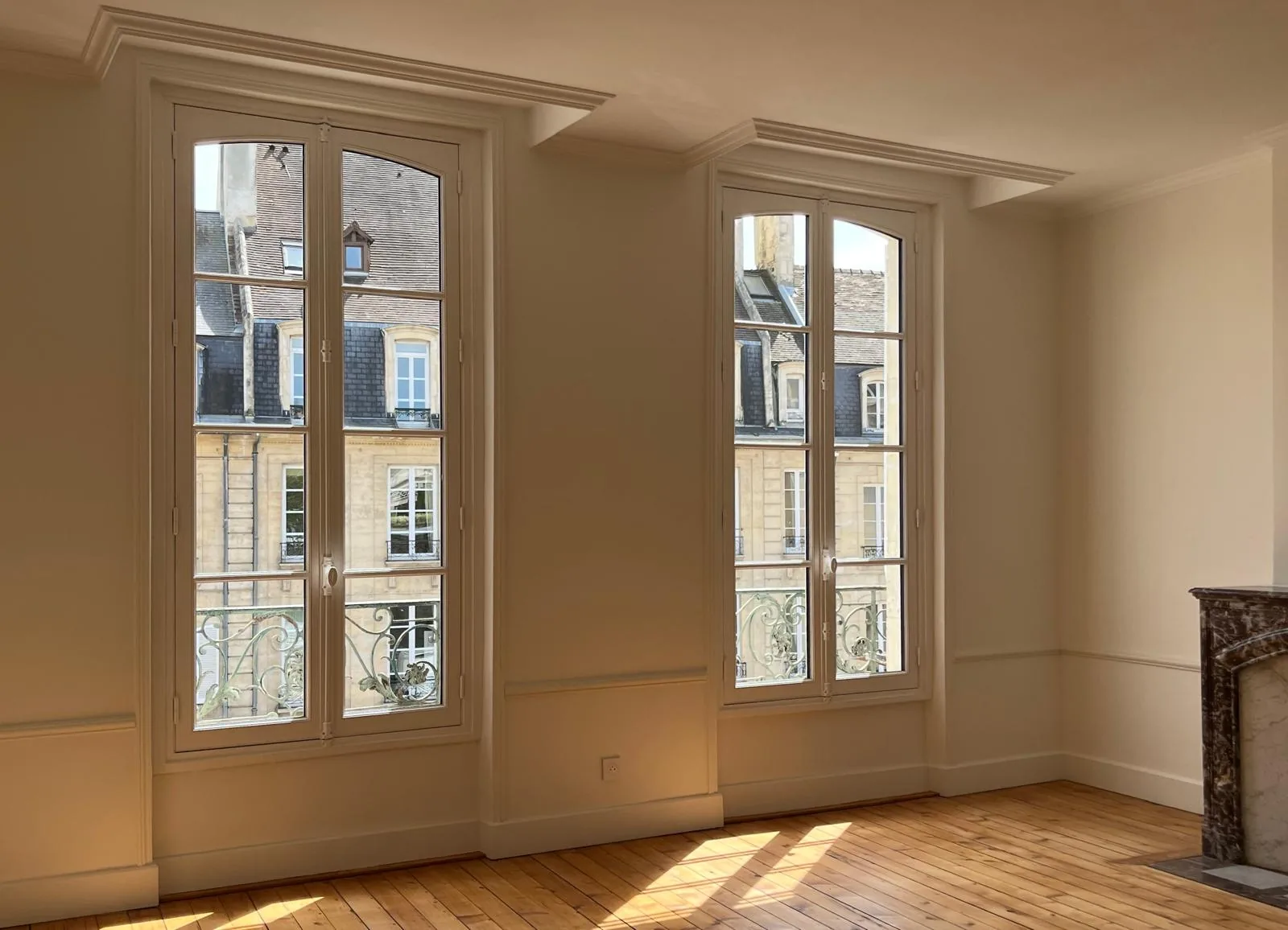 Deux hautes fenêtres bordées de moulures dans une pièce aux murs clairs et au parquet bois.