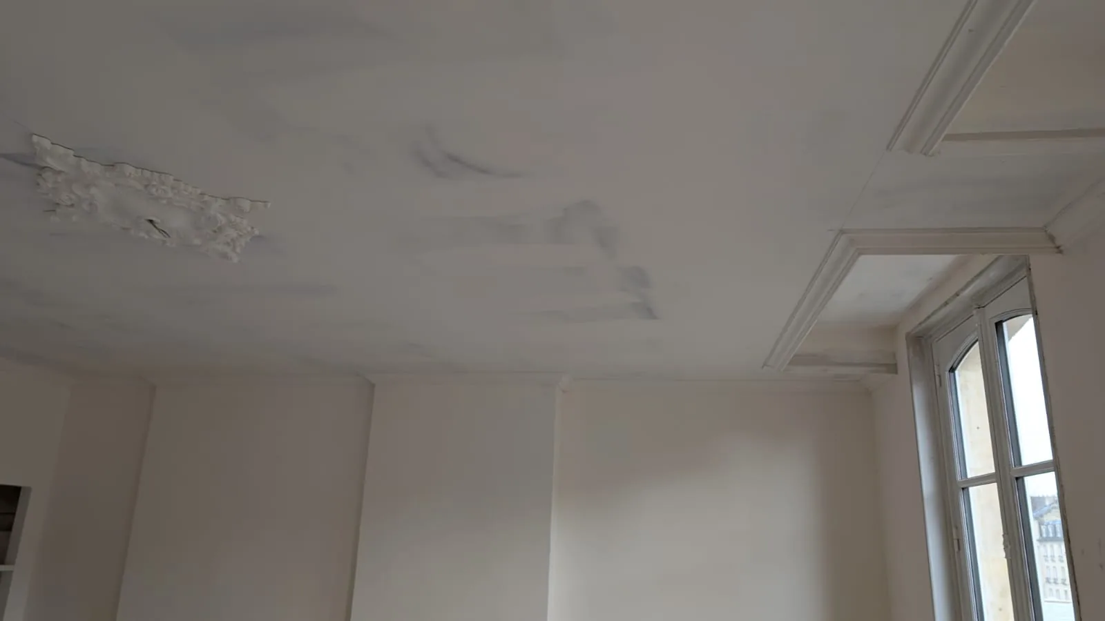 Corniches et ornements de plafond anciens partiellement poncés et enduits autour d’une fenêtre.