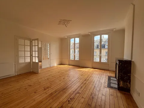 Grande pièce vide avec rosace, corniches et moulures murales blanches, parquet bois et cheminée en marbre.